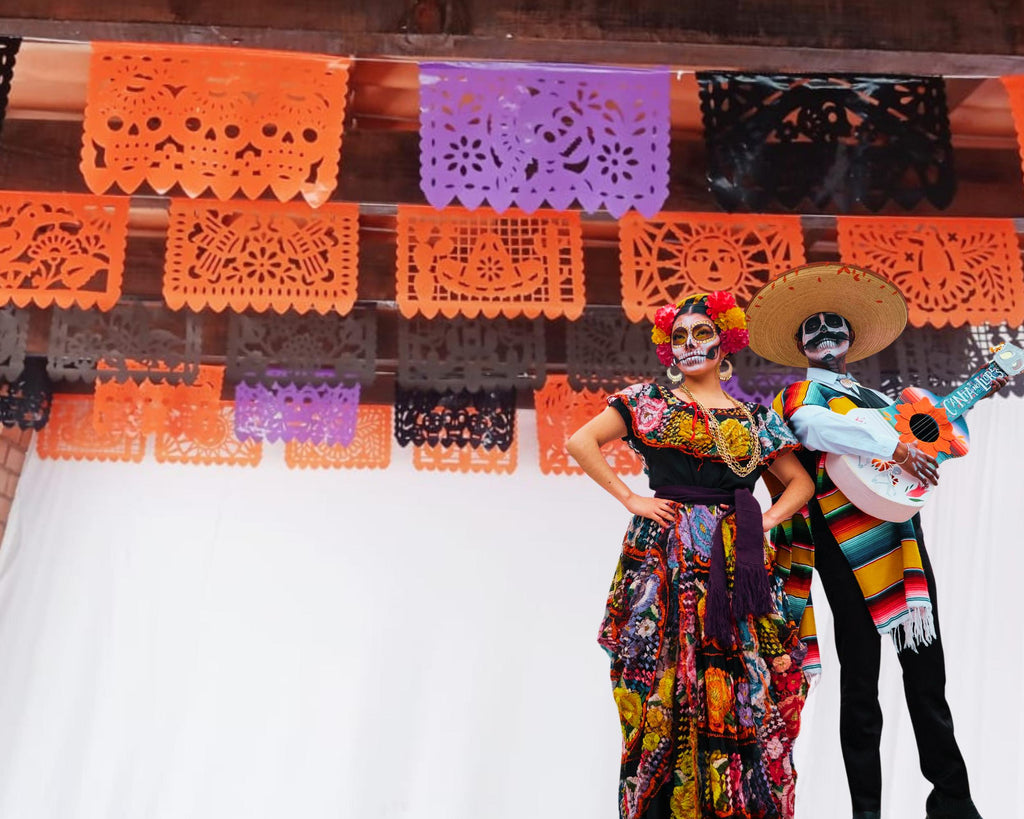 Two people in colorful costumes with a decorative background featuring papel picado.