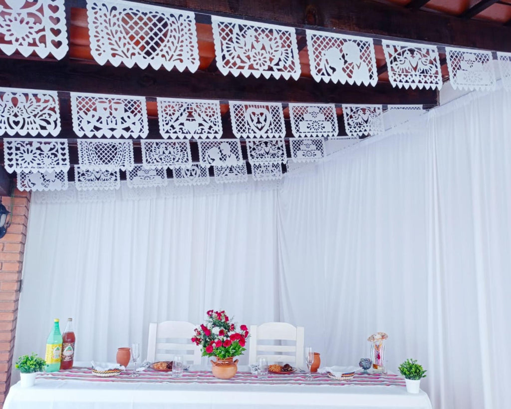 Decorative white papel picado banner above a table set with flowers and food.