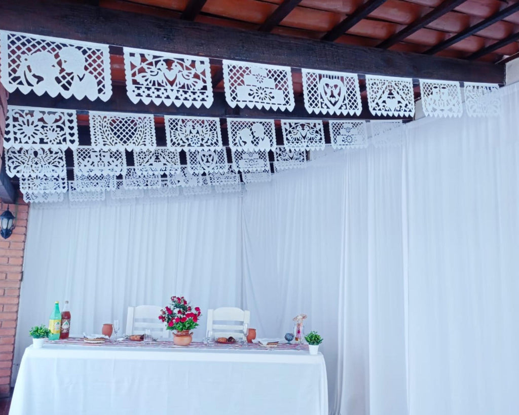 Decorative papel picado above a table with flowers and candles on a white curtain background.