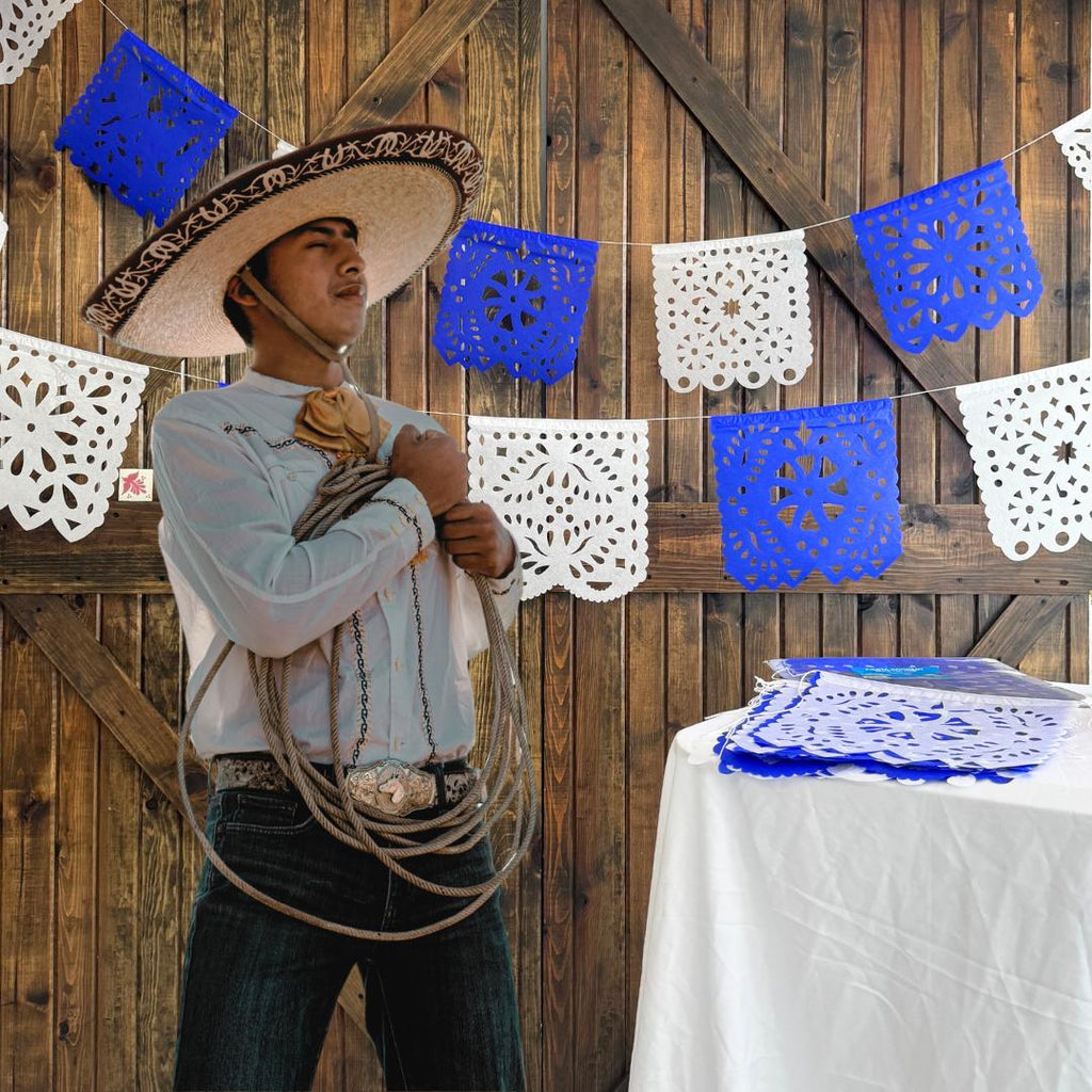 Person in cowboy hat with lasso standing against a wooden wall with blue and white papel picado.