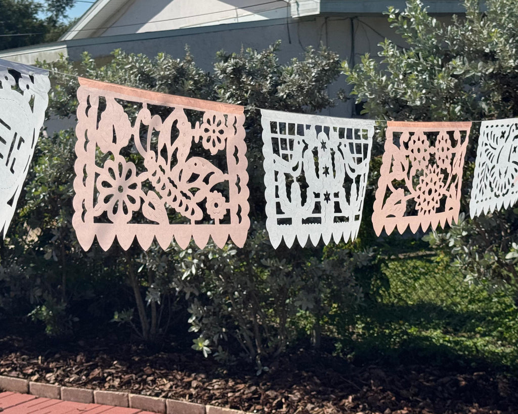 Decorative papel picado banners hanging on a fence with greenery in the background.